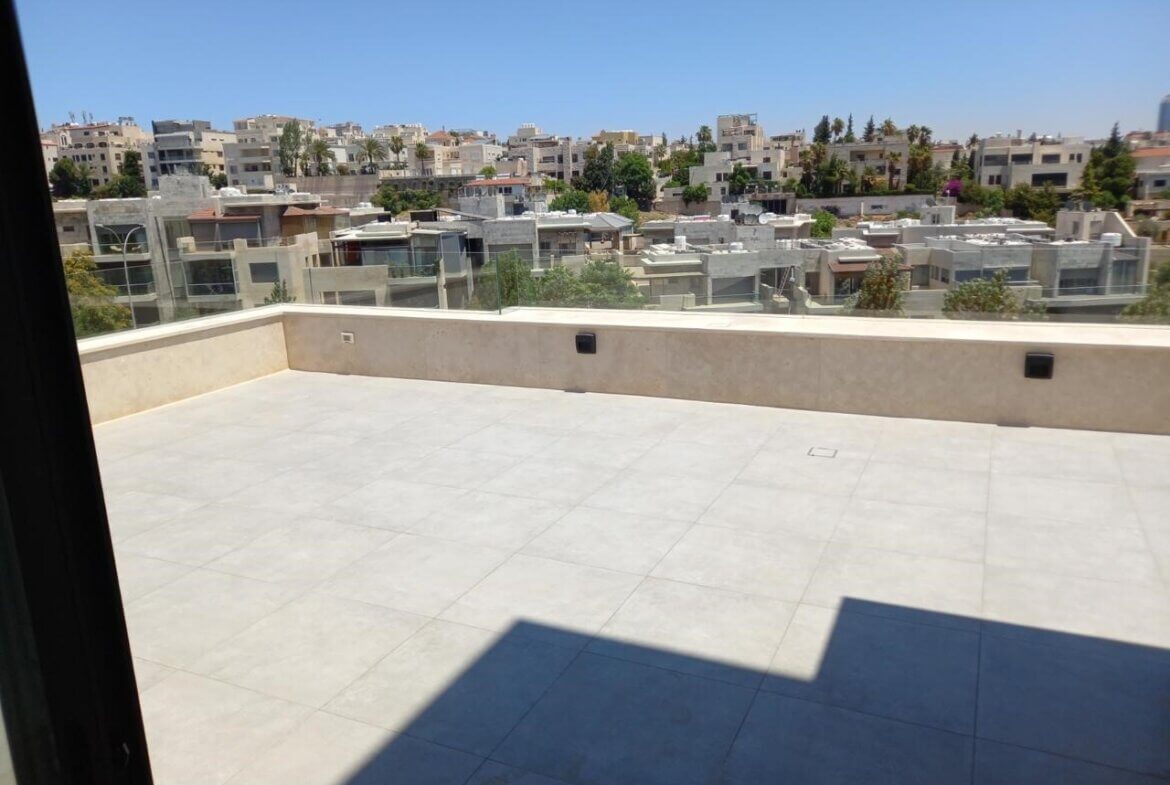 777491c4-d255-4cf4-80f9-5ae1c9027ba3 Sunny rooftop terrace with light tile floor and beige parapet overlooking a dense cityscape of low-rise buildings and palm trees.