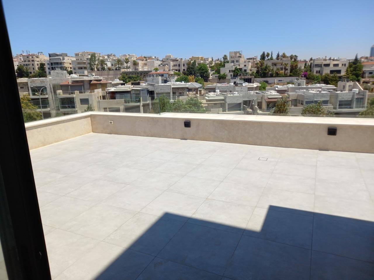 777491c4-d255-4cf4-80f9-5ae1c9027ba3 Sunny rooftop terrace with light tile floor and beige parapet overlooking a dense cityscape of low-rise buildings and palm trees.