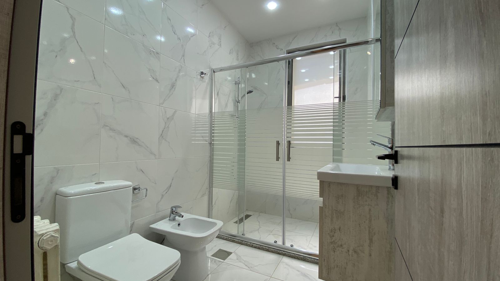 787b6c9b-f9ce-426d-8fd4-3792ab1d64dc Modern white bathroom with marble tiles, toilet, bidet, and a glass-walled shower enclosure with frosted stripes and chrome fittings