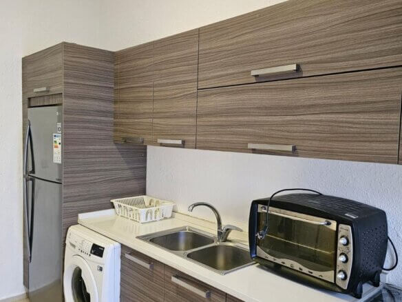 Compact kitchen-laundry area with a tall stainless refrigerator, front-loading washing machine, double sink, dish rack, and wood-grain cabinets above and below the counter.