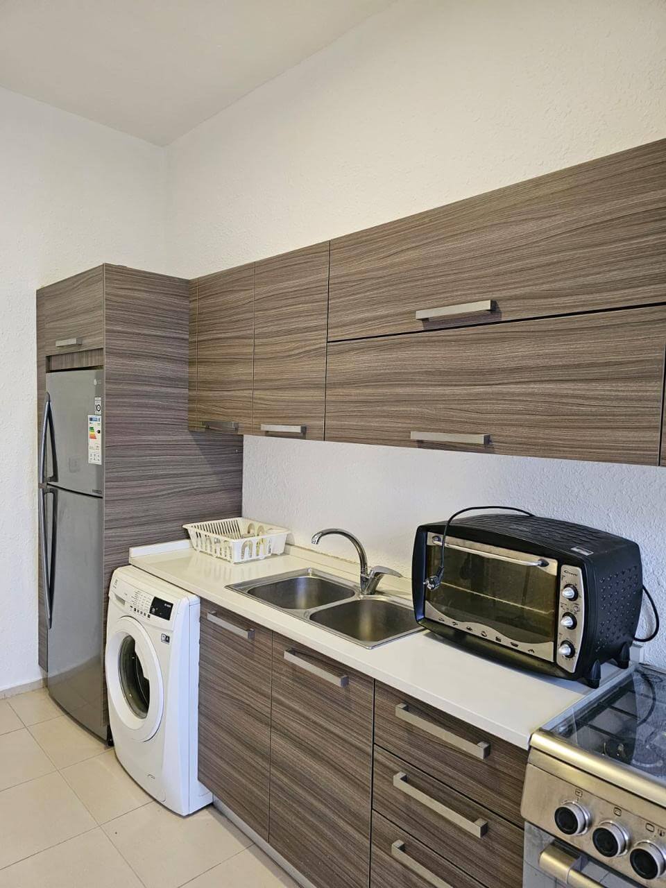 78953742-05ba-4b87-886d-362e28b159bf Compact kitchen-laundry area with a tall stainless refrigerator, front-loading washing machine, double sink, dish rack, and wood-grain cabinets above and below the counter.