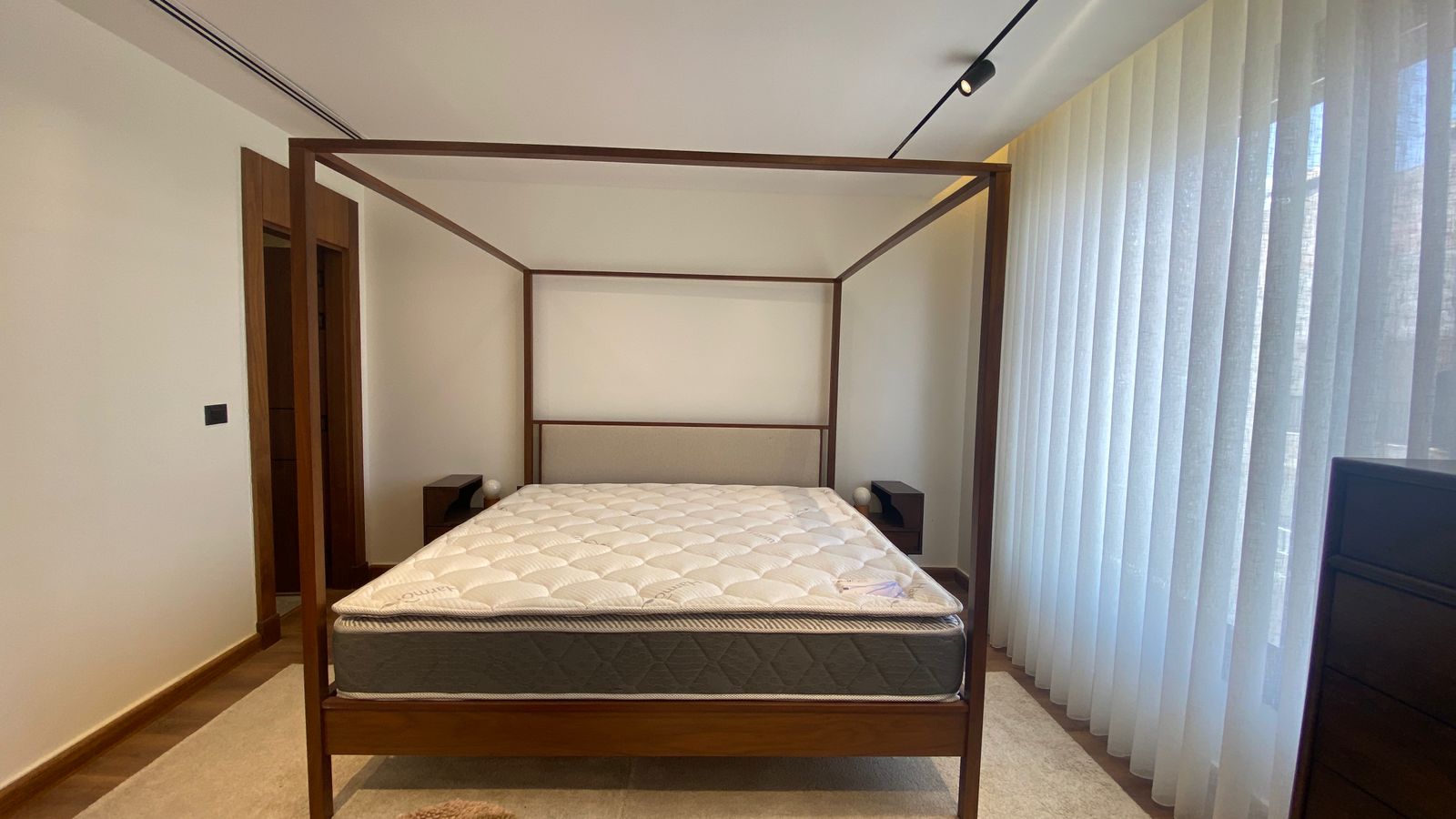 7920e6cf-386f-4761-8fd5-f2f0c6df4790 Wood four-poster bed with mattress in a minimalist bedroom, flanked by dark nightstands on each side