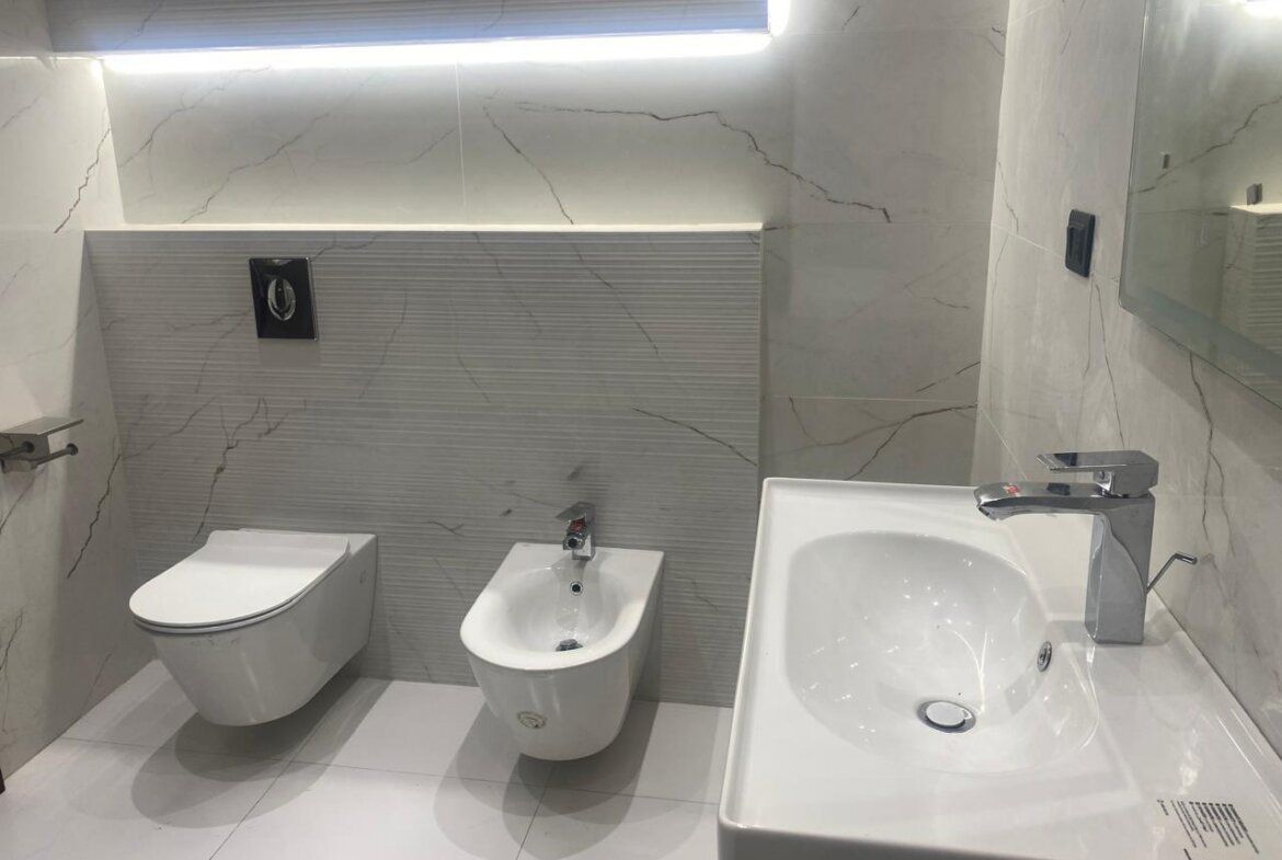 Modern white bathroom with a wall-mounted toilet and bidet beside a white sink on marble walls and floor.