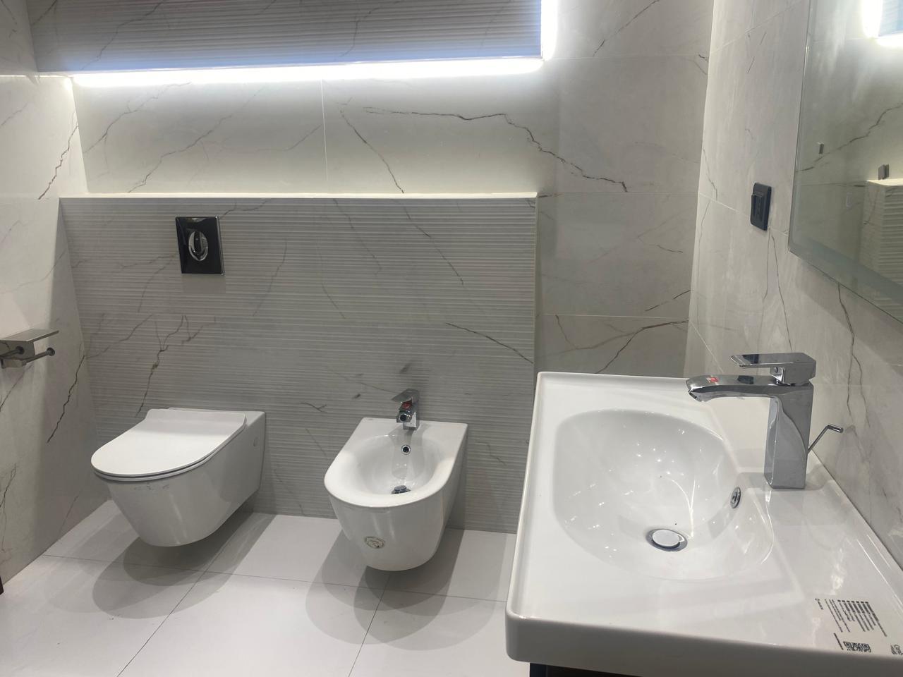 Modern white bathroom with a wall-mounted toilet and bidet beside a white sink on marble walls and floor.