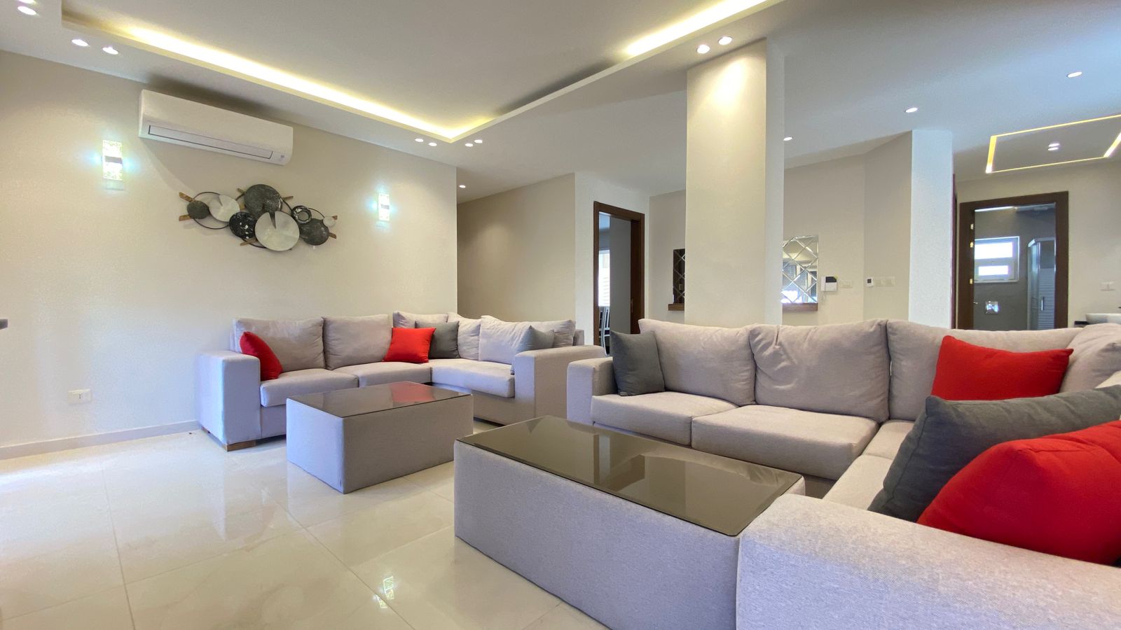 793d2da6-a9e8-4c45-aa29-3538445eba75 Bright living room with an L-shaped gray sofa, red and gray cushions, two glass-topped coffee tables, and wall art under warm ceiling lighting.
