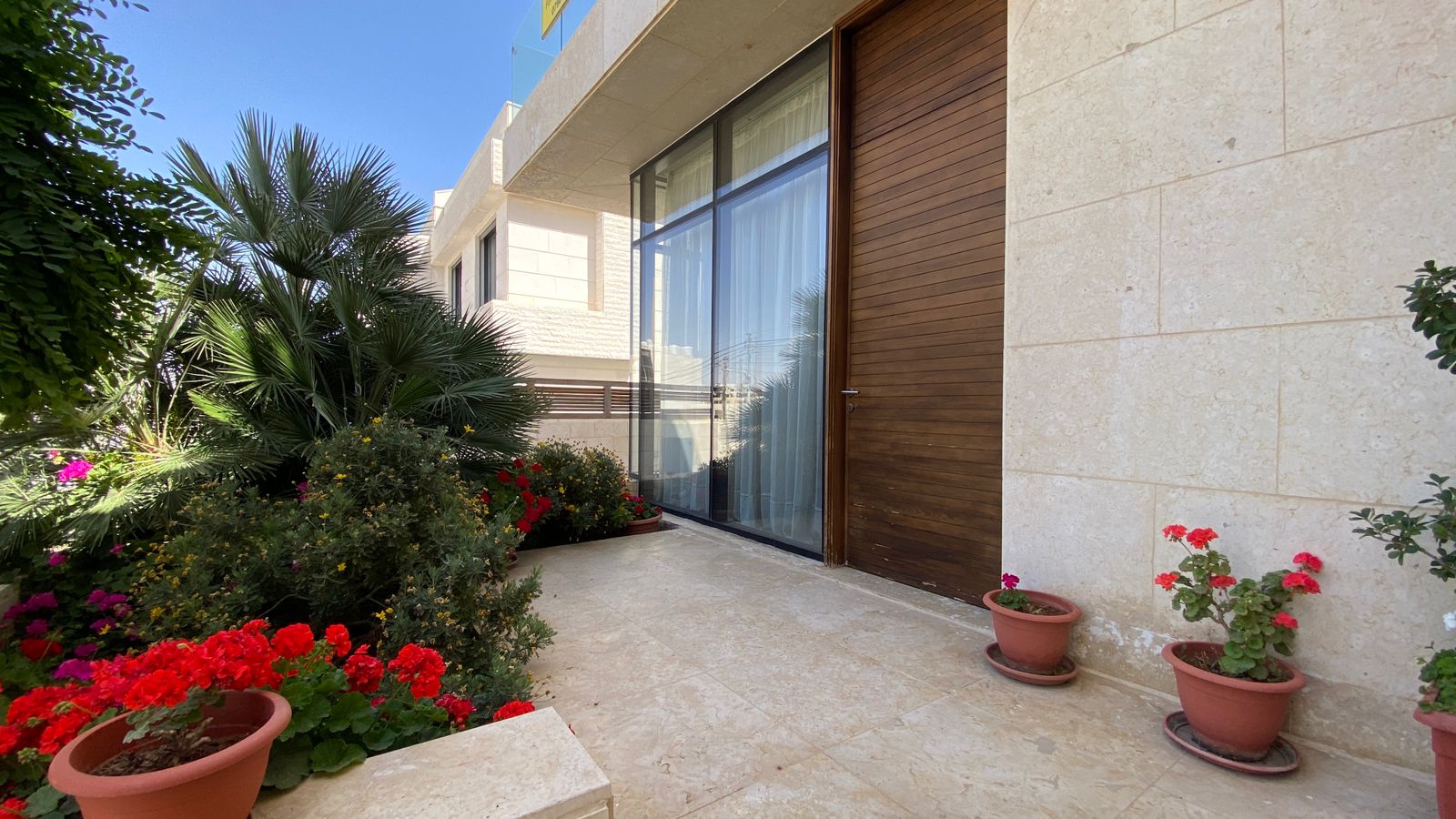 79e9554b-88b6-4d0b-9c67-91ce6855b2f8 Entrance to a modern home with a glass sliding door and a wooden door, flanked by potted red geraniums and lush greenery.
