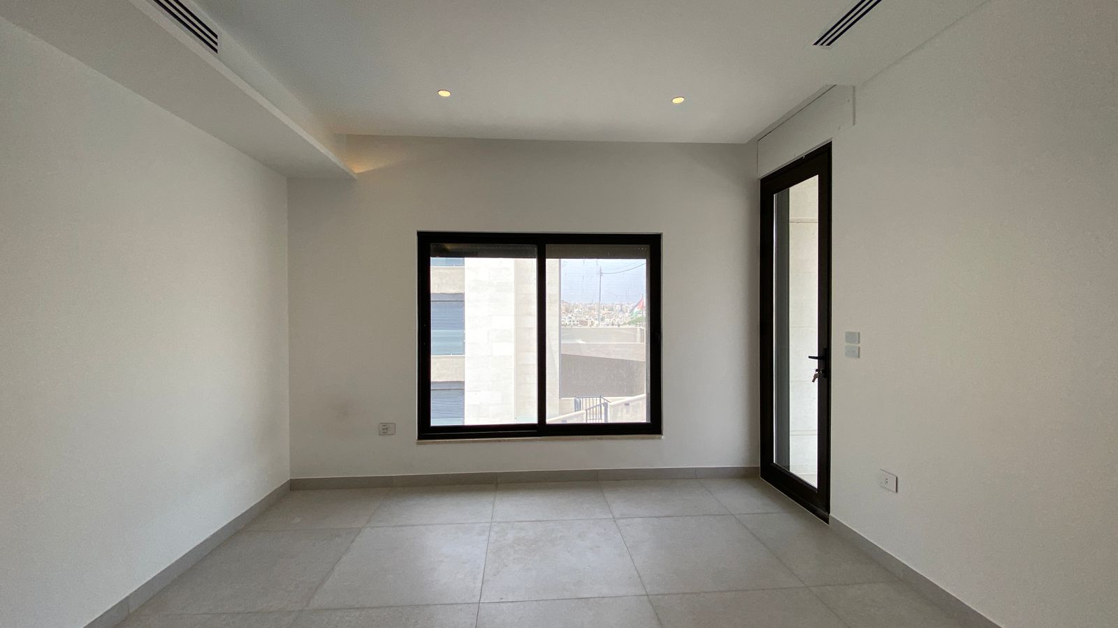 79fa15c3-12a0-48d0-ae49-fba371492bbd Empty, unfurnished room with white walls, gray tiled floor, a large central window, and a glass door to a balcony on the right; ceiling has recessed lights and vents.
