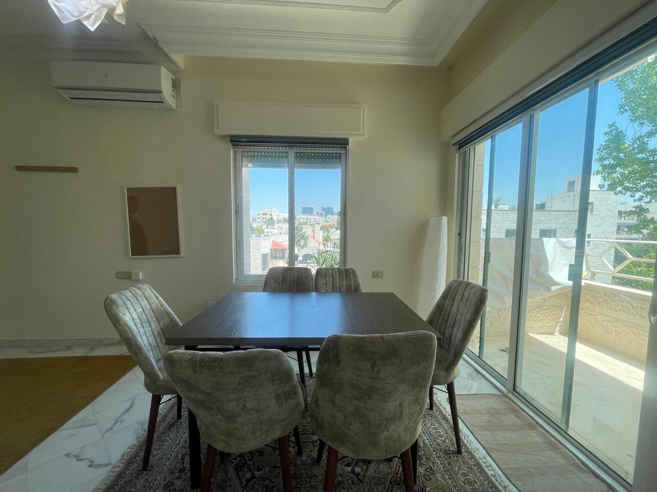 7a0135e5-498c-49ed-beb0-4f420b836a6f Bright dining room with a dark wood table and six gray upholstered chairs, marble floor, and large sliding glass doors to a balcony with a city view outside.