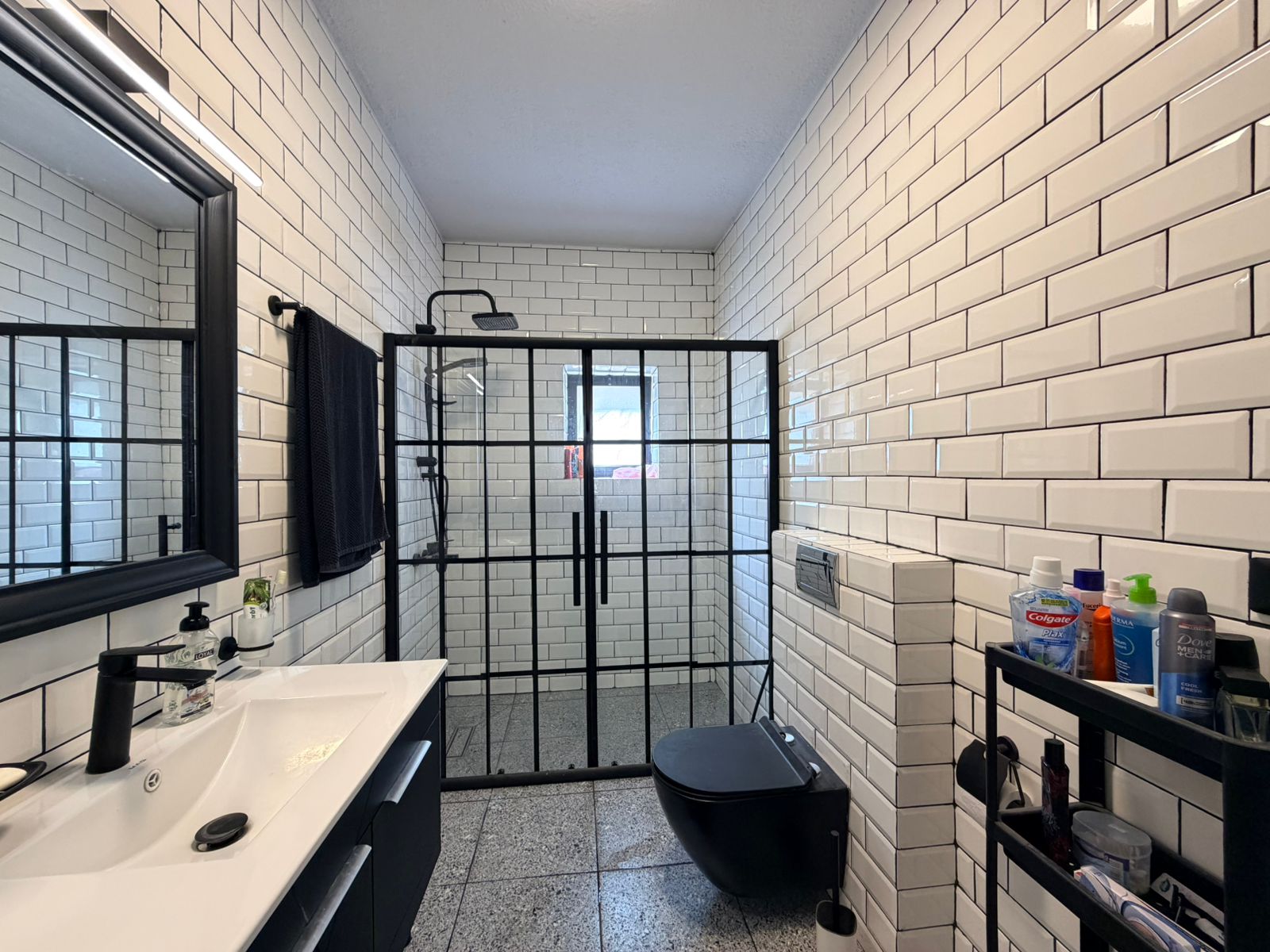 7a234f5c-abea-4410-a04b-06e9b0b7e615 Modern bathroom with white subway tile walls, black-framed glass shower, and a double vanity with a white sink and black faucet.