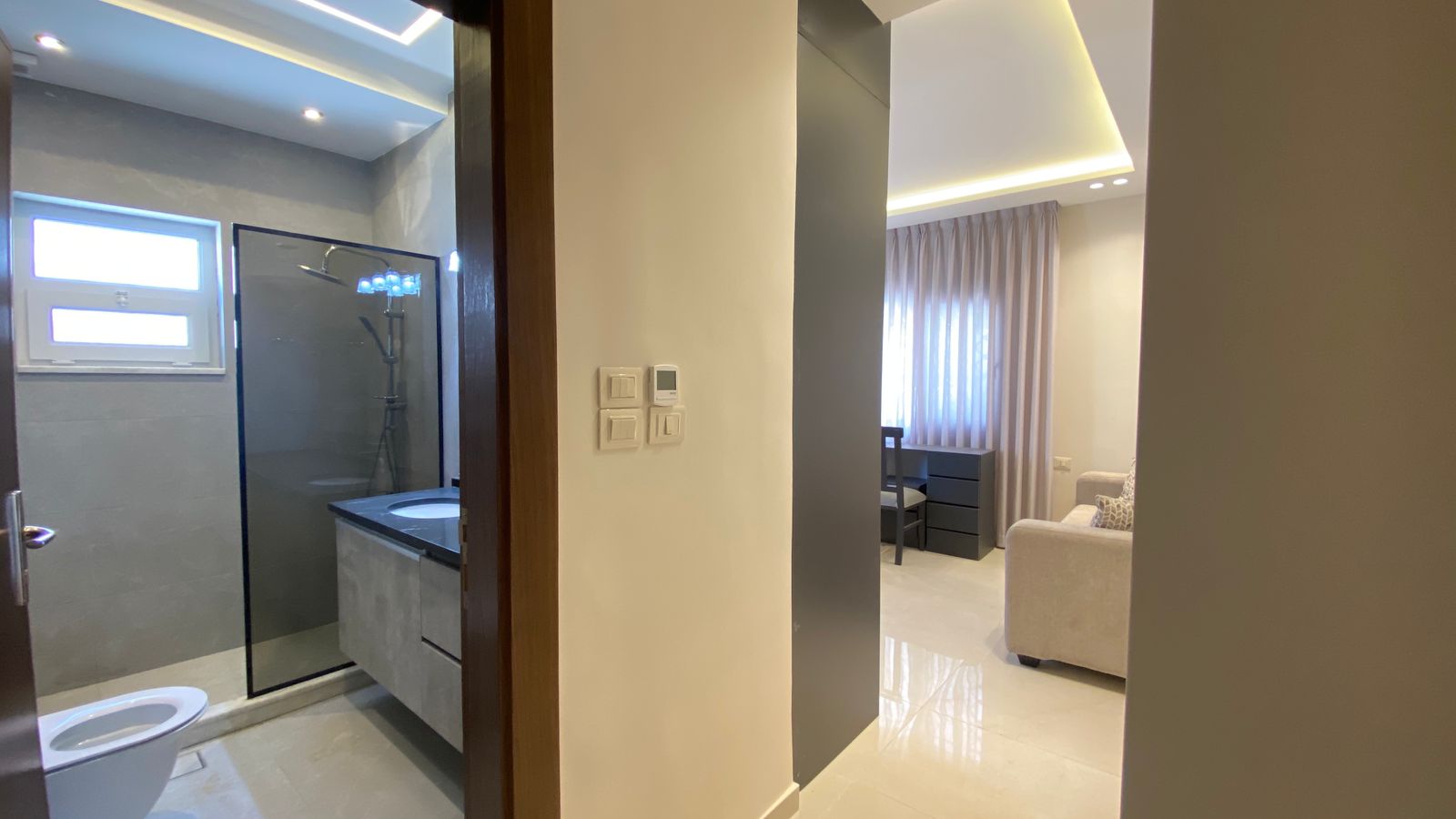 7bd4ddf2-1002-431d-9f80-48bac940a487 Modern bathroom with glass shower enclosure and vanity on left; hallway opens to a living area with a beige sofa and desk under sheer curtains.