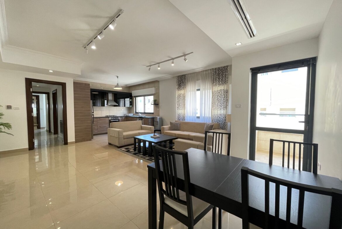 Open-concept living space with beige sofas around a dark coffee table, adjacent dining table, and a modern kitchen in the background.