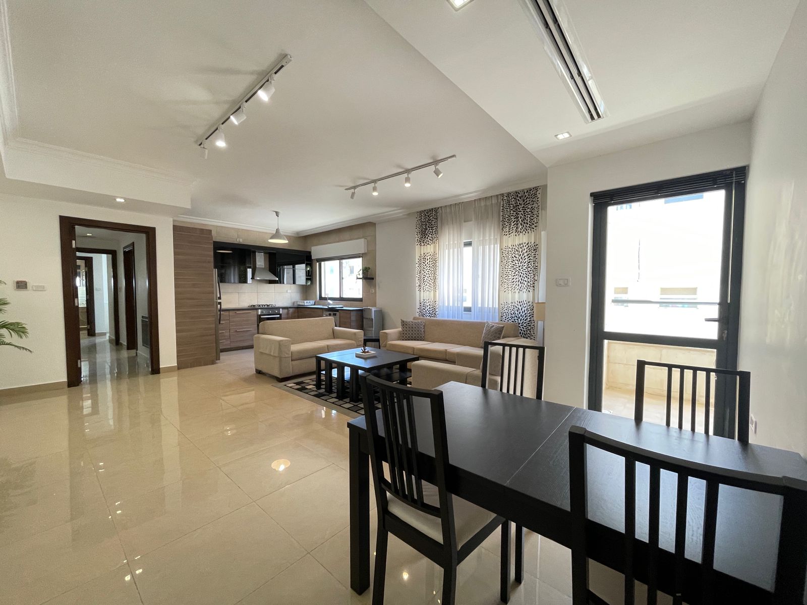 7c1e4423-67f5-4b93-a072-9e587b3b8bd7 Open-concept living space with beige sofas around a dark coffee table, adjacent dining table, and a modern kitchen in the background.
