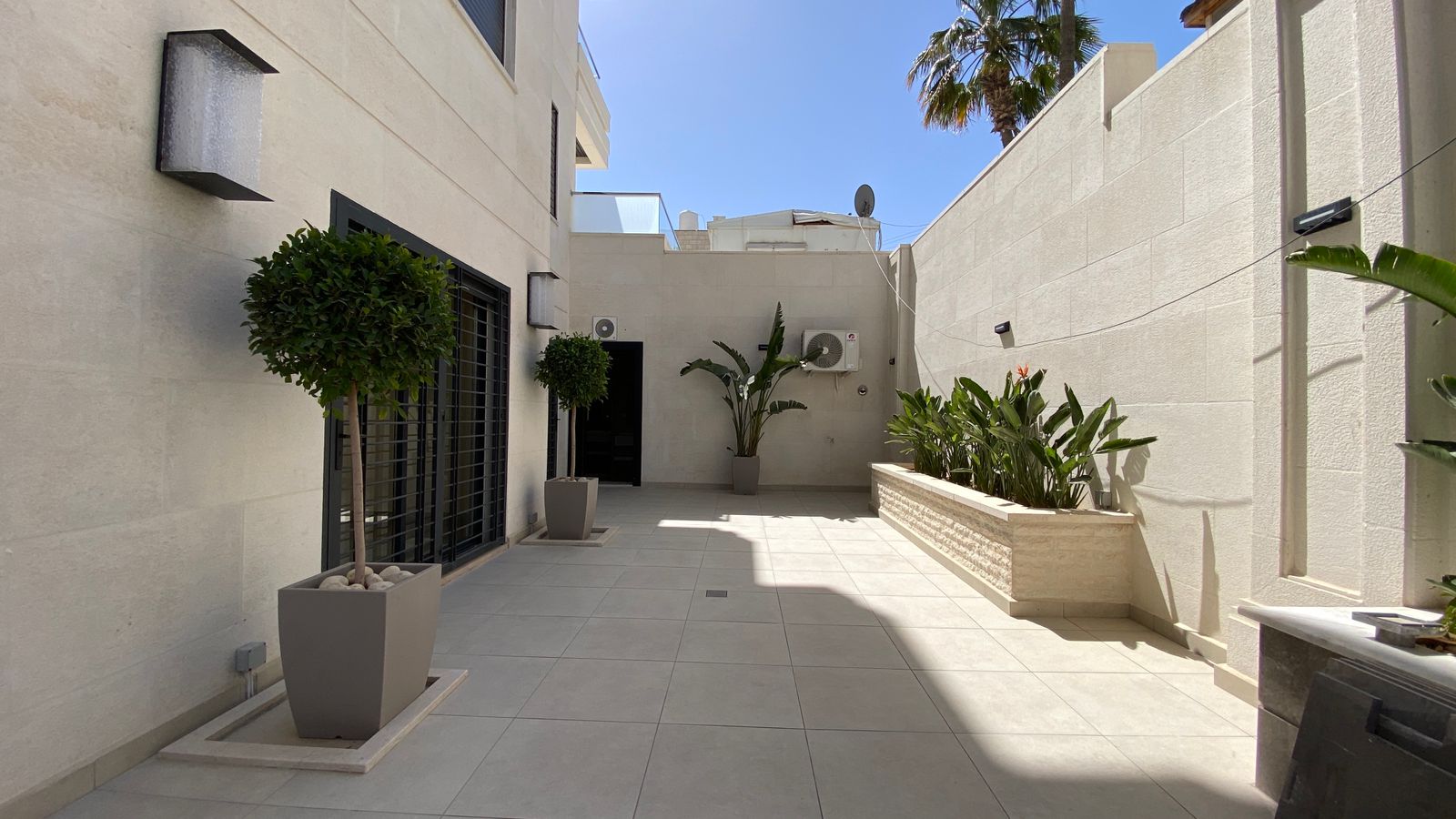 7cf3c62e-fc3b-4d2f-9475-3892b3a7bba6 Bright modern courtyard with beige walls, large tiled floor, and potted topiary trees along the left and back wall.