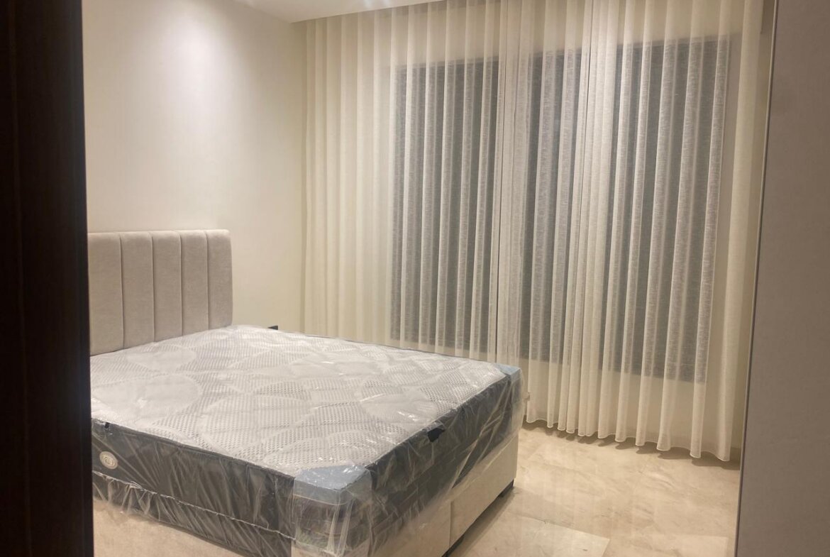 Queen-size bed with a padded beige headboard in a minimalist bedroom; mattress still in plastic wrap on a light-colored frame/a bed waiting to be used.