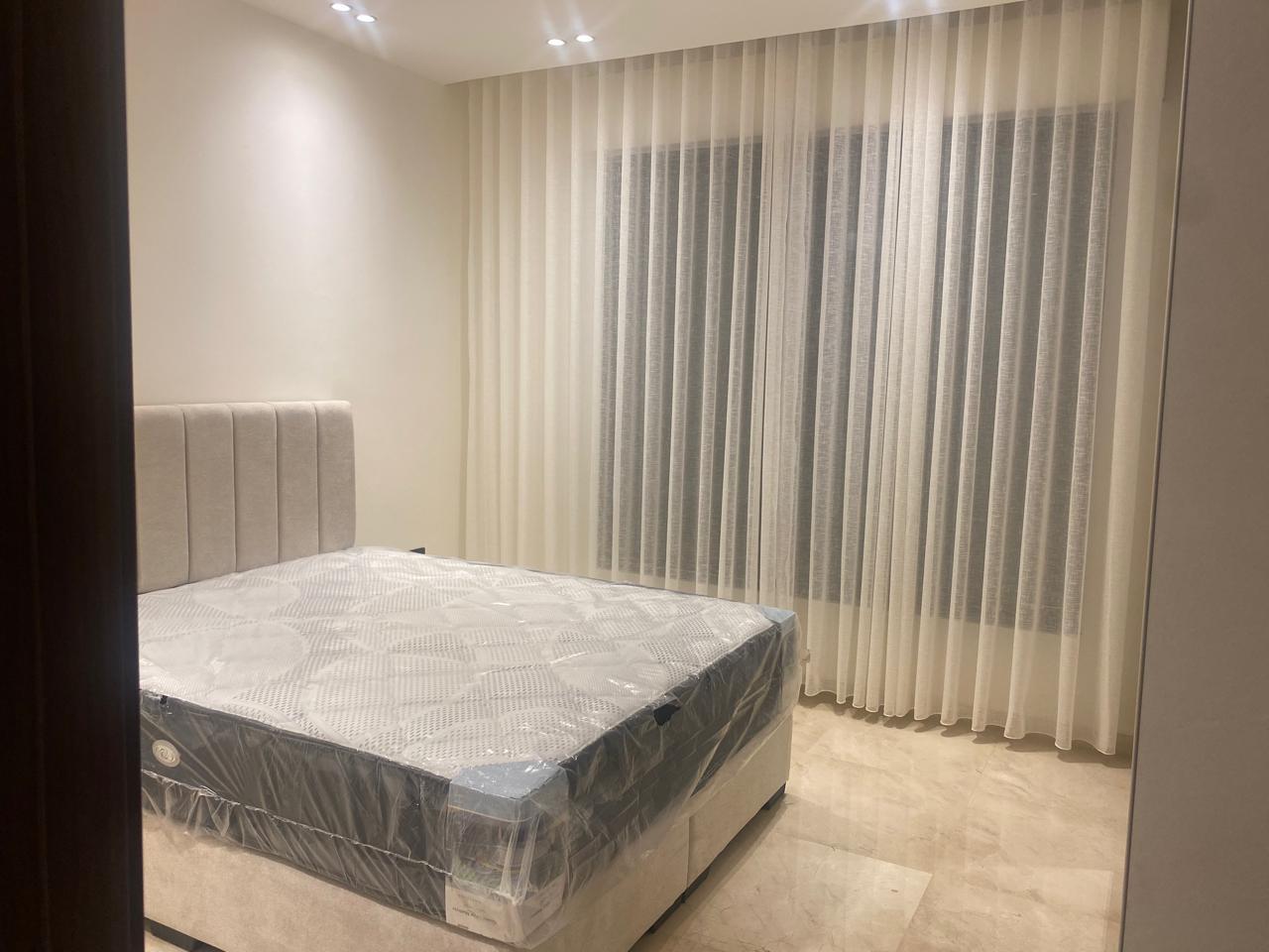 Queen-size bed with a padded beige headboard in a minimalist bedroom; mattress still in plastic wrap on a light-colored frame/a bed waiting to be used.