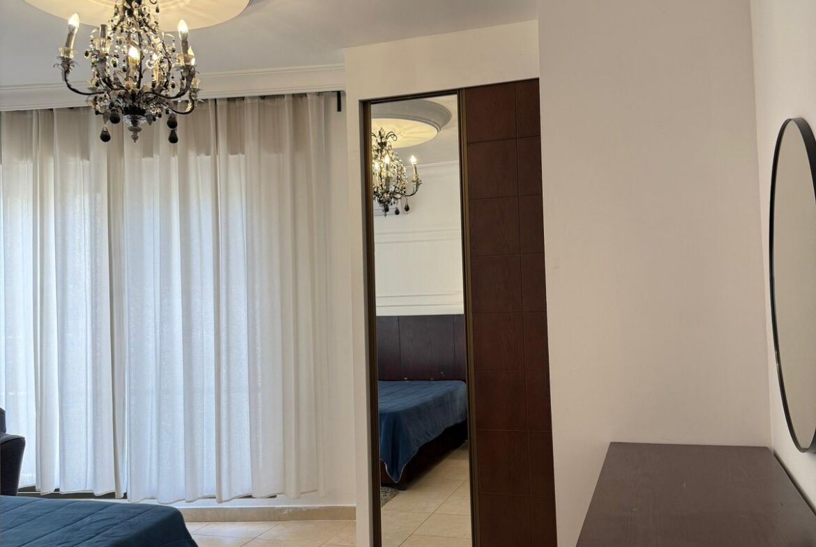 Bedroom with a decorative chandelier hanging from a round ceiling medallion, white sheer curtains, and a blue-bedspread visible by a mirrored closet door.
