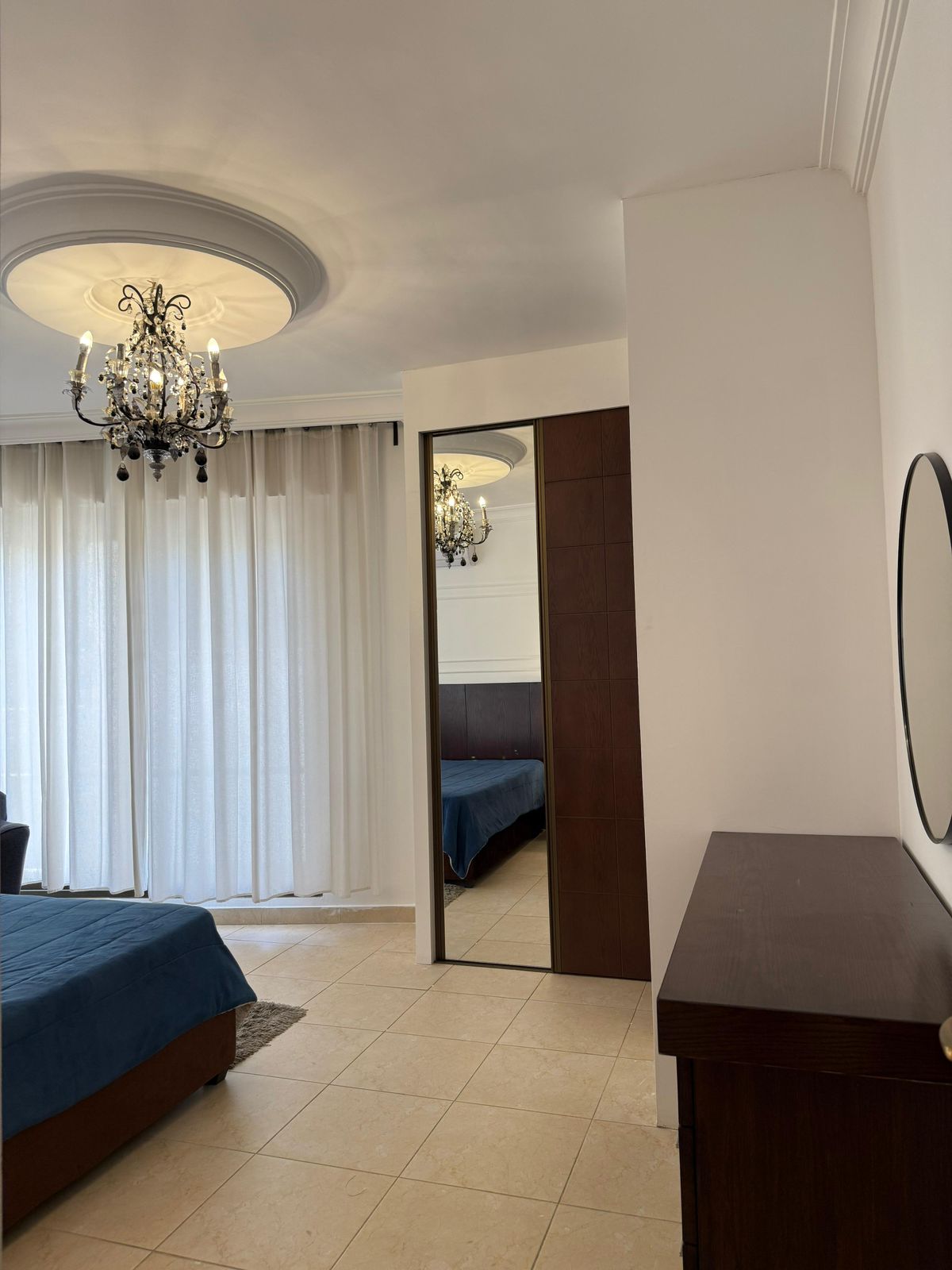 Bedroom with a decorative chandelier hanging from a round ceiling medallion, white sheer curtains, and a blue-bedspread visible by a mirrored closet door.