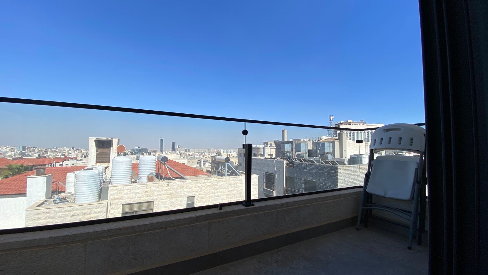 800acc21-5d7a-4db9-84cf-b557b5292f5d Balcony with a clear blue sky overlooking a city skyline and rooftop water tanks; a folded chair rests on the right edge.