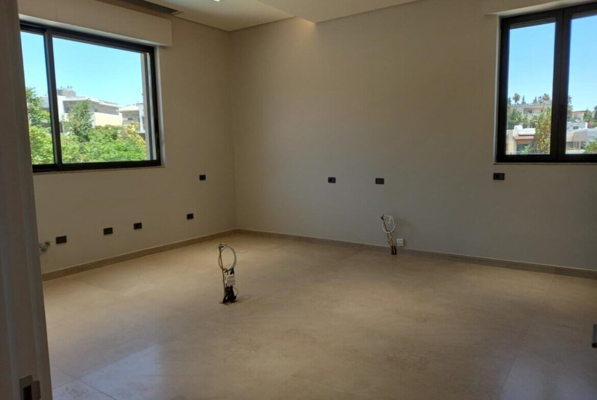 800bee2d-364a-4ea4-bfb9-5492ba309b31 Empty, unfurnished room under construction with two large windows and visible electrical outlets/wires on walls and floor.