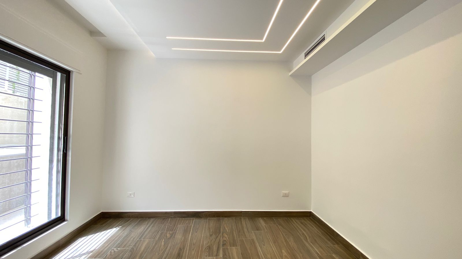 80922c4b-e885-4a60-9b76-57b5cf721aee Empty modern room with white walls, wood-look flooring, large left window with blinds, and recessed linear LED ceiling lights.