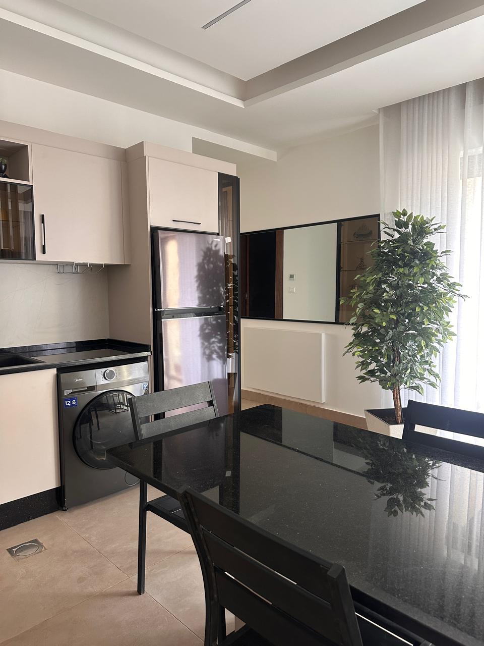 80e00e94-9ef4-47a5-b7b2-24323506832c Modern kitchen with beige cabinets, stainless fridge, washing machine, and a glossy black dining table with chairs.