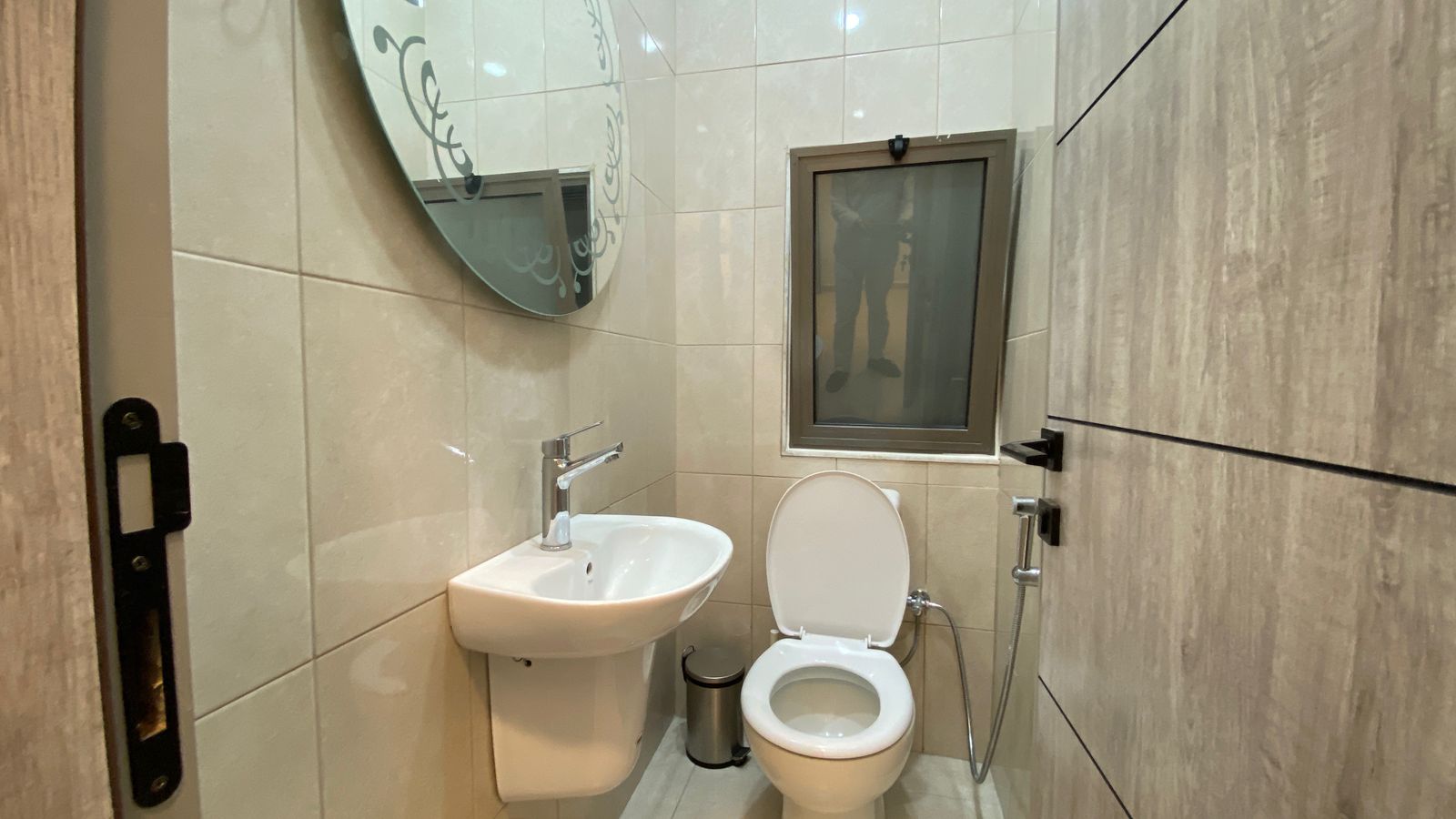 810f2270-4268-403c-a1ef-43a62c57848c Small bathroom with wall-mounted sink, toilet with open lid, and a handheld bidet sprayer by the windowed wall.