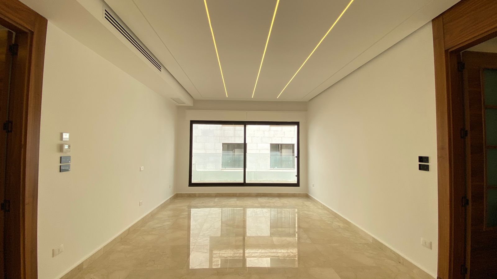 8117c2ea-2a6d-4ef6-97cb-716249255502 Bright empty living room with a large window at the far end, marble floor and recessed linear LED ceiling lights across the ceiling.