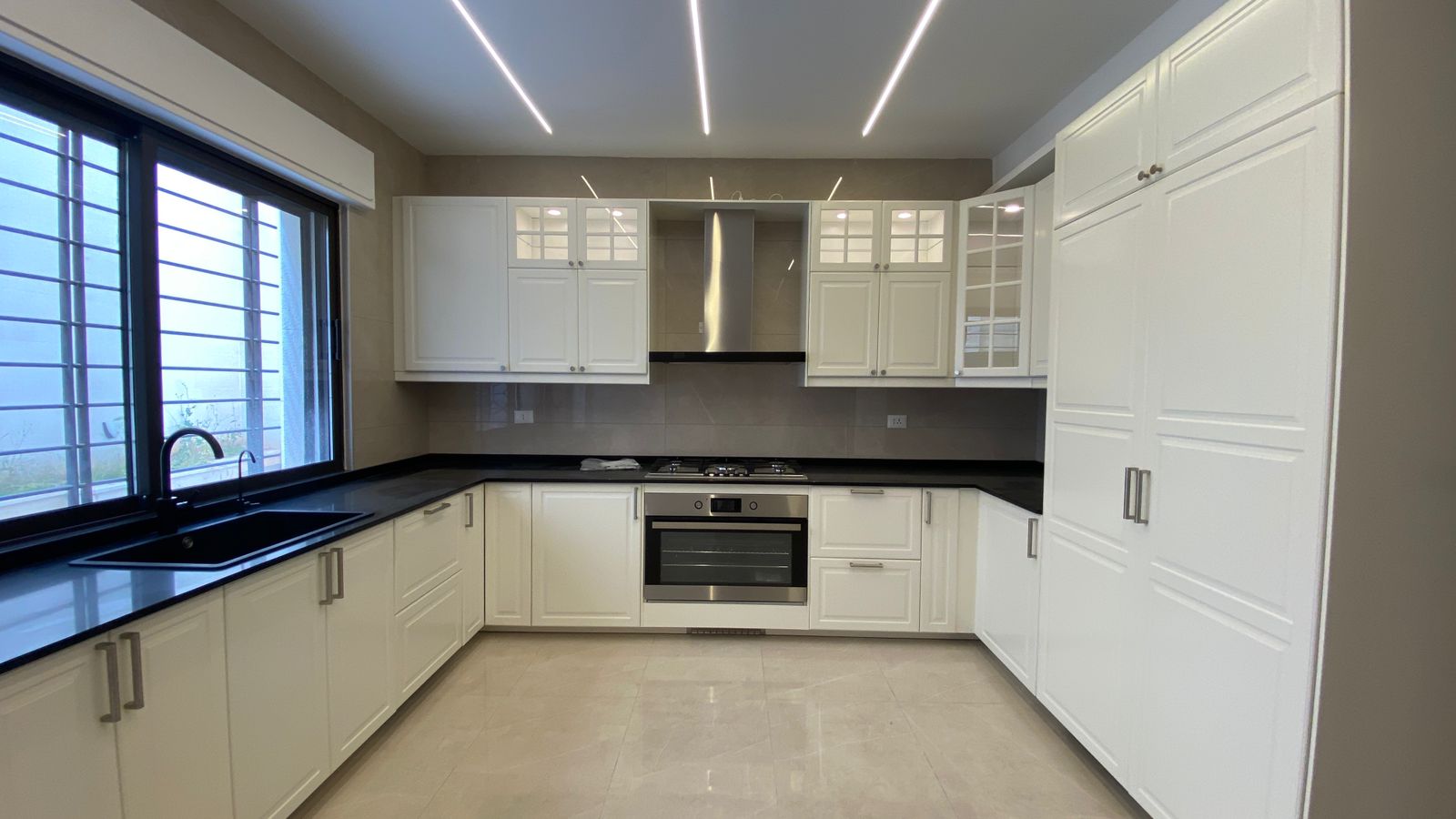 8117fd9b-bd33-4751-a858-8de489dc0472 Bright white U-shaped kitchen with black countertops, stainless steel oven and range hood, and a large window with blinds on the left.