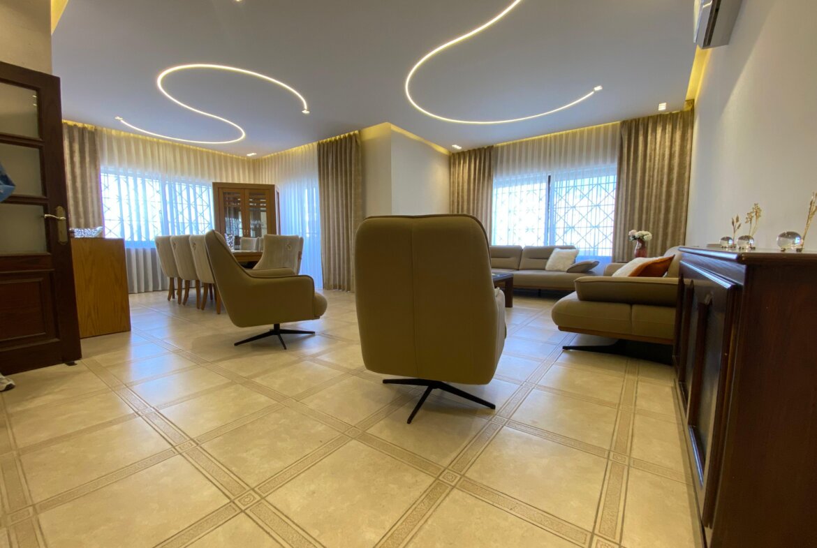 Spacious living room with beige recliners, a beige sofa, and a wooden cabinet under large windows with sheer curtains; curved ceiling lights stretch overhead.