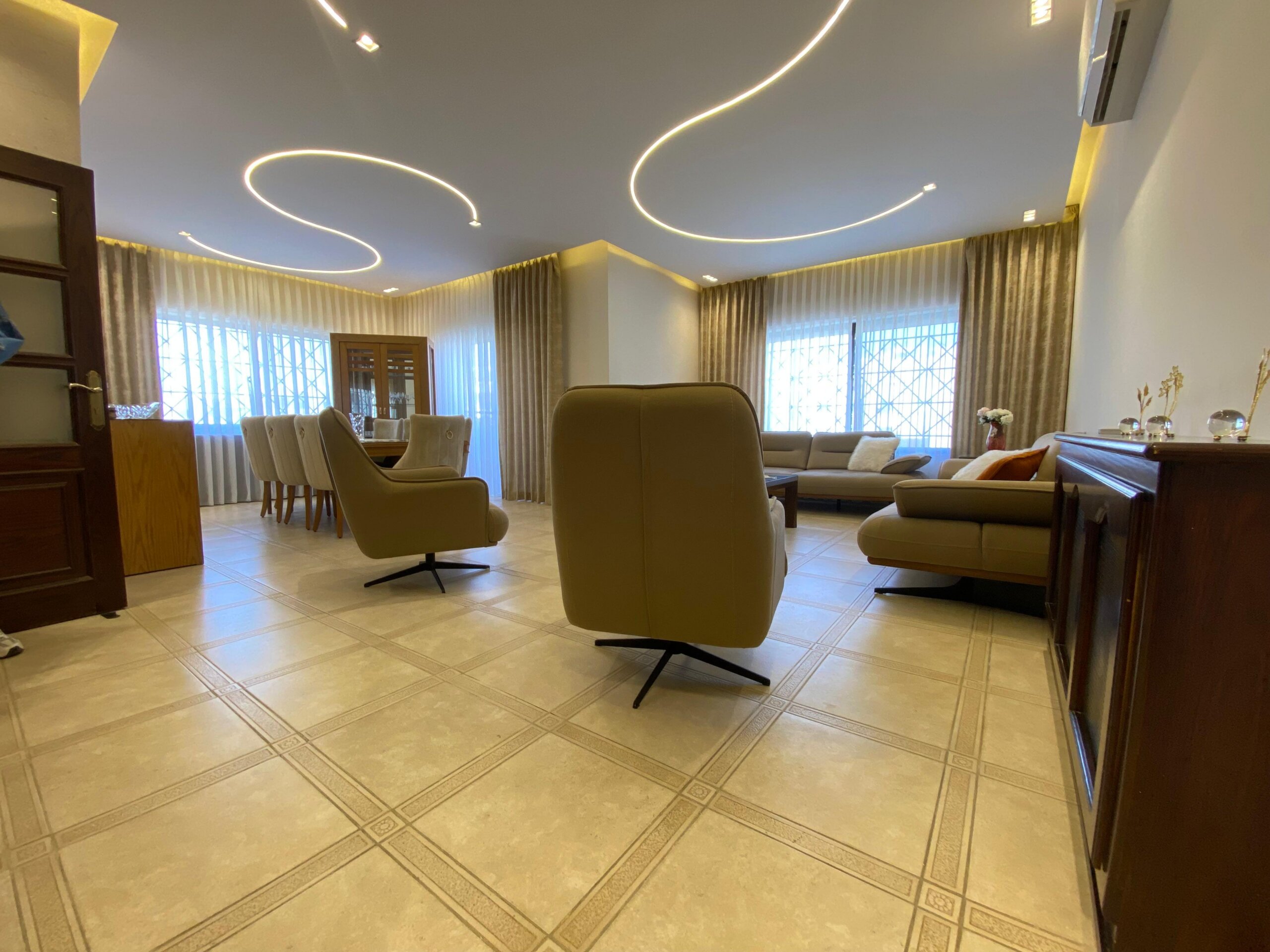 81218aa7-5346-4d90-835d-4beae8e1c5af Spacious living room with beige recliners, a beige sofa, and a wooden cabinet under large windows with sheer curtains; curved ceiling lights stretch overhead.