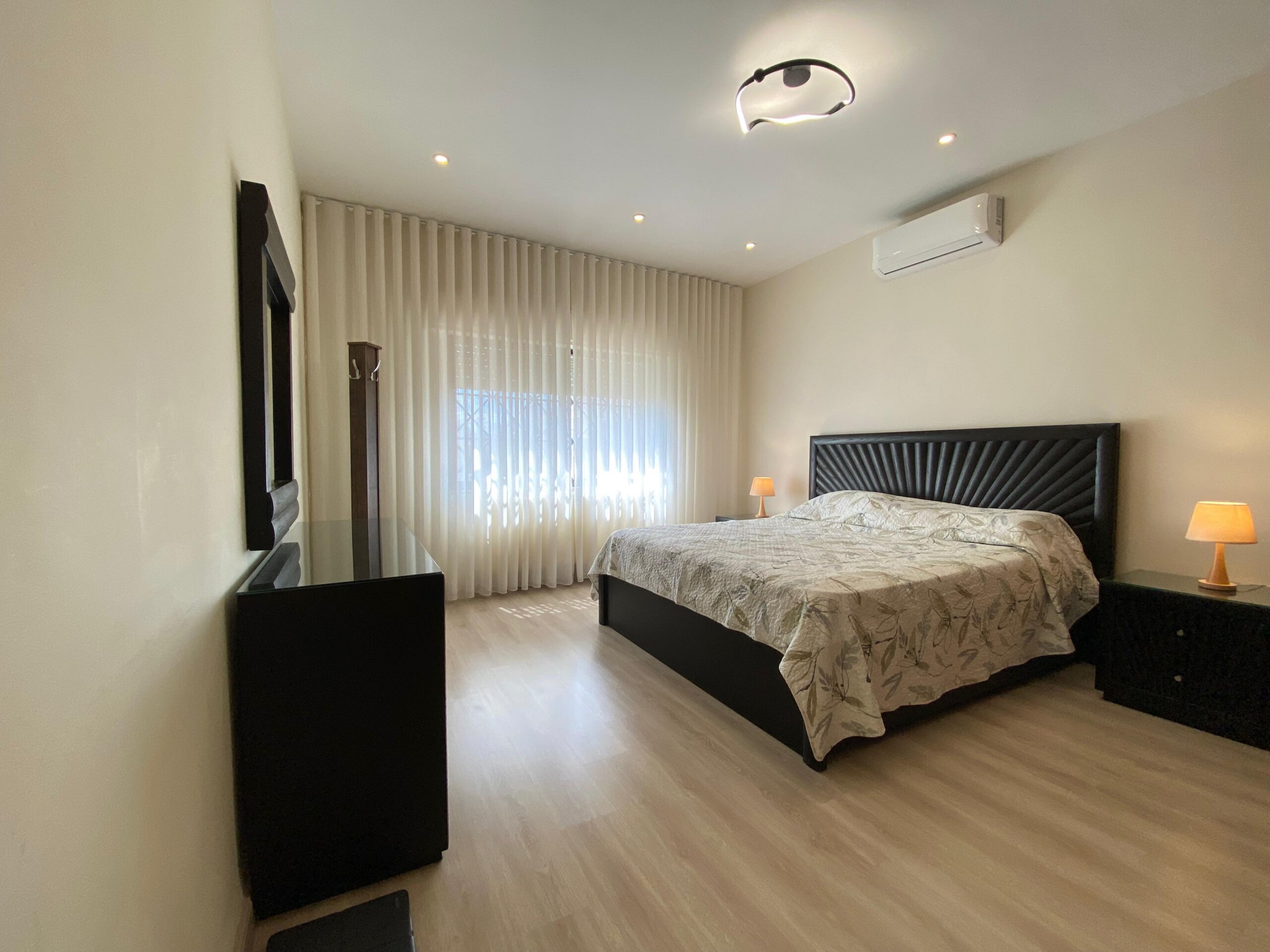 823581f7-d9b7-4c22-9d6e-cfff90b69ca8 Bright hotel-style bedroom with two large beds, dark wood headboards, and matching nightstands beneath soft ceiling lighting and sheer curtains. An air conditioner is mounted high on the wall.