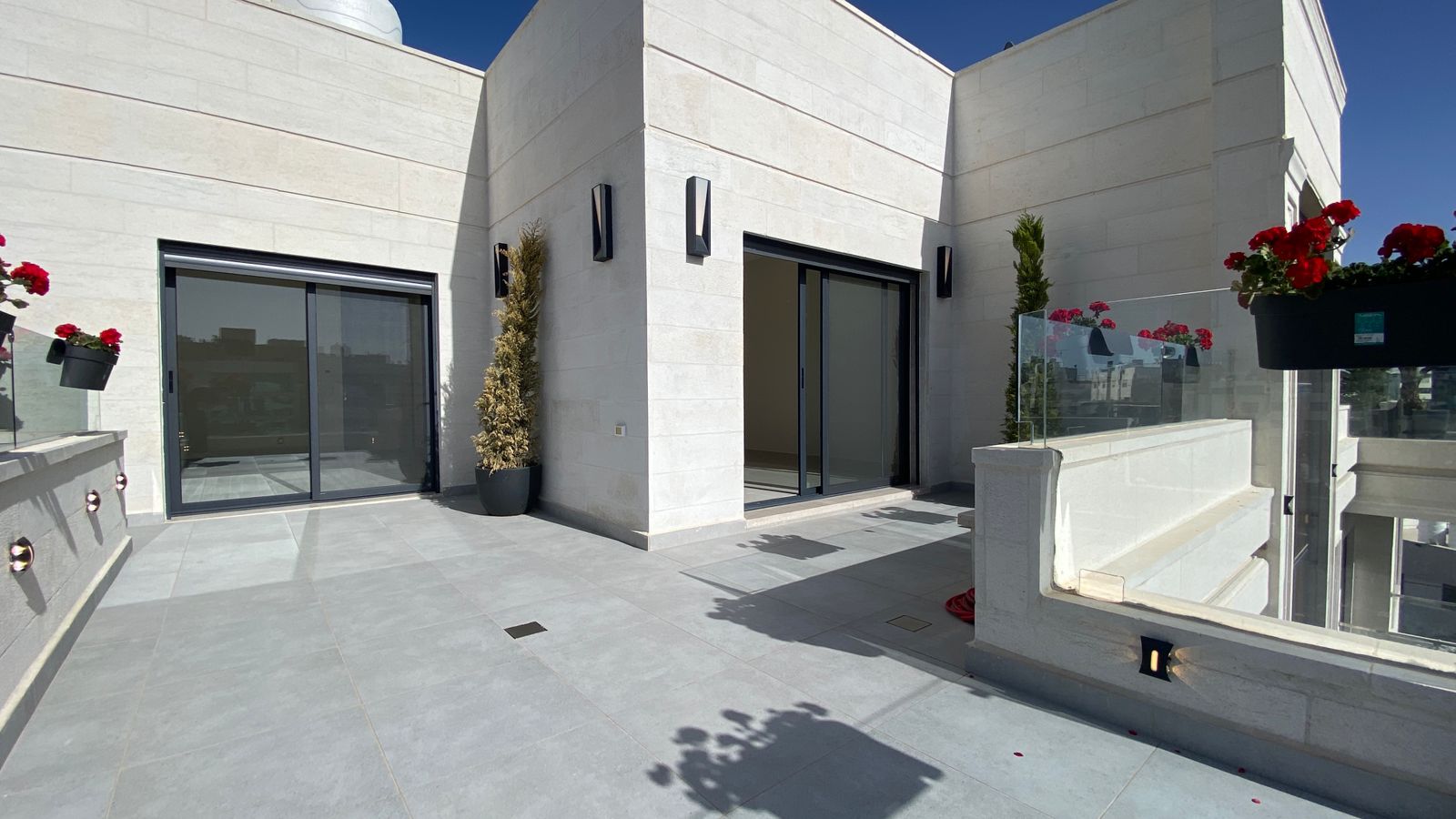 837955a0-2ea5-4f24-abd8-2a22d4452a33 Modern white stone terrace with two sliding glass doors, potted plants, and a glass railing on a sunny day.