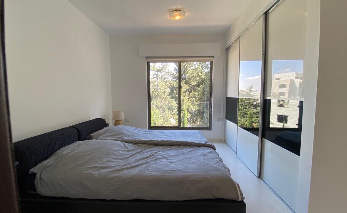 84e49adf-62a5-4614-a2ec-13f04e794e6c Modern bedroom with a low platform bed, gray bedding, and a small bedside lamp; large window shows trees outside.
