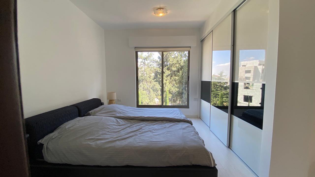 84e49adf-62a5-4614-a2ec-13f04e794e6c Modern bedroom with a low platform bed, gray bedding, and a small bedside lamp; large window shows trees outside.