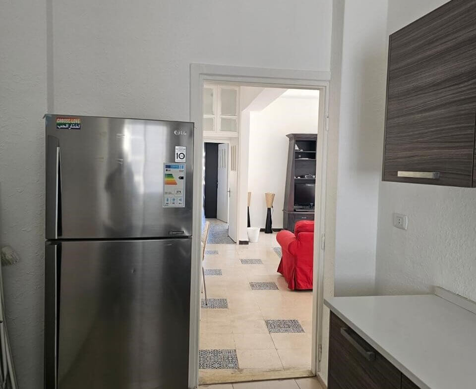 8511ed15-e876-4a15-81f7-56d1e2b83c42 Stainless steel fridge in a bright kitchen with dark wood cabinets and a doorway to a living room with a red sofa.