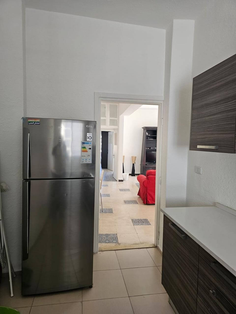 8511ed15-e876-4a15-81f7-56d1e2b83c42 Stainless steel fridge in a bright kitchen with dark wood cabinets and a doorway to a living room with a red sofa.