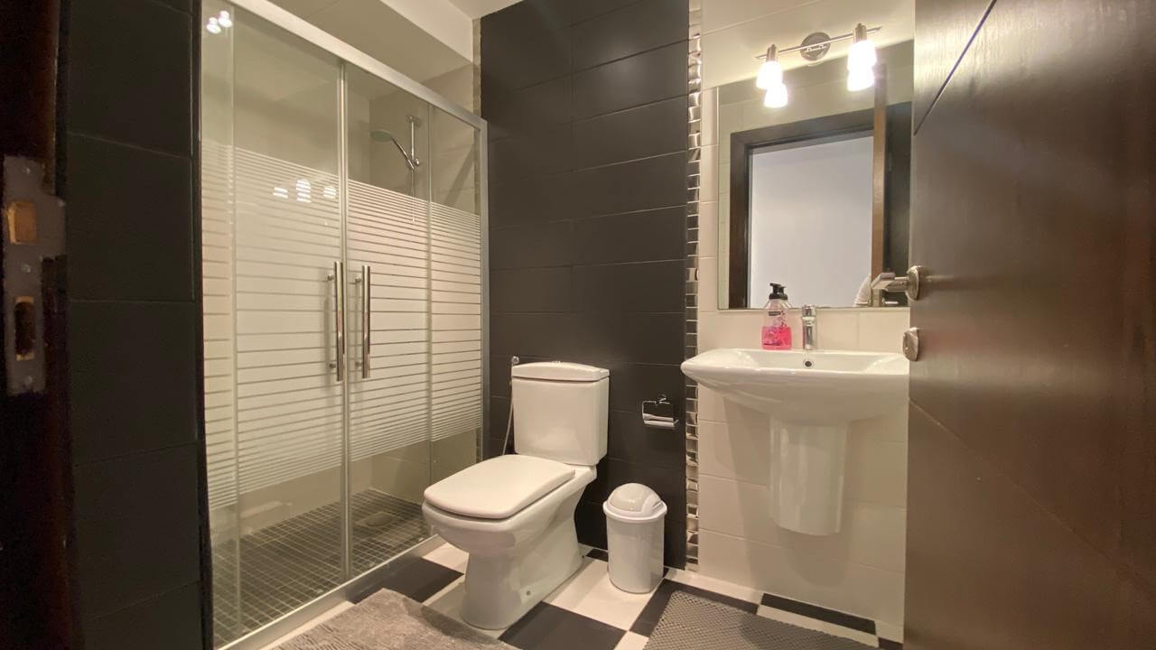 85f91377-2d40-410f-8878-afe30fb13e37 Modern bathroom with glass shower enclosure, white toilet, and pedestal sink; white tile walls with dark accents and a mirror above the sink.