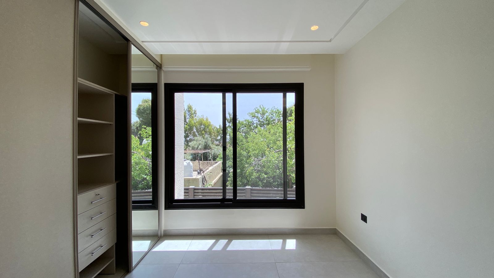 868680b7-b1de-4c5e-bb5a-f295e602d680 Empty modern bedroom with built-in wardrobe on the left and a large black-framed window showing green trees outside