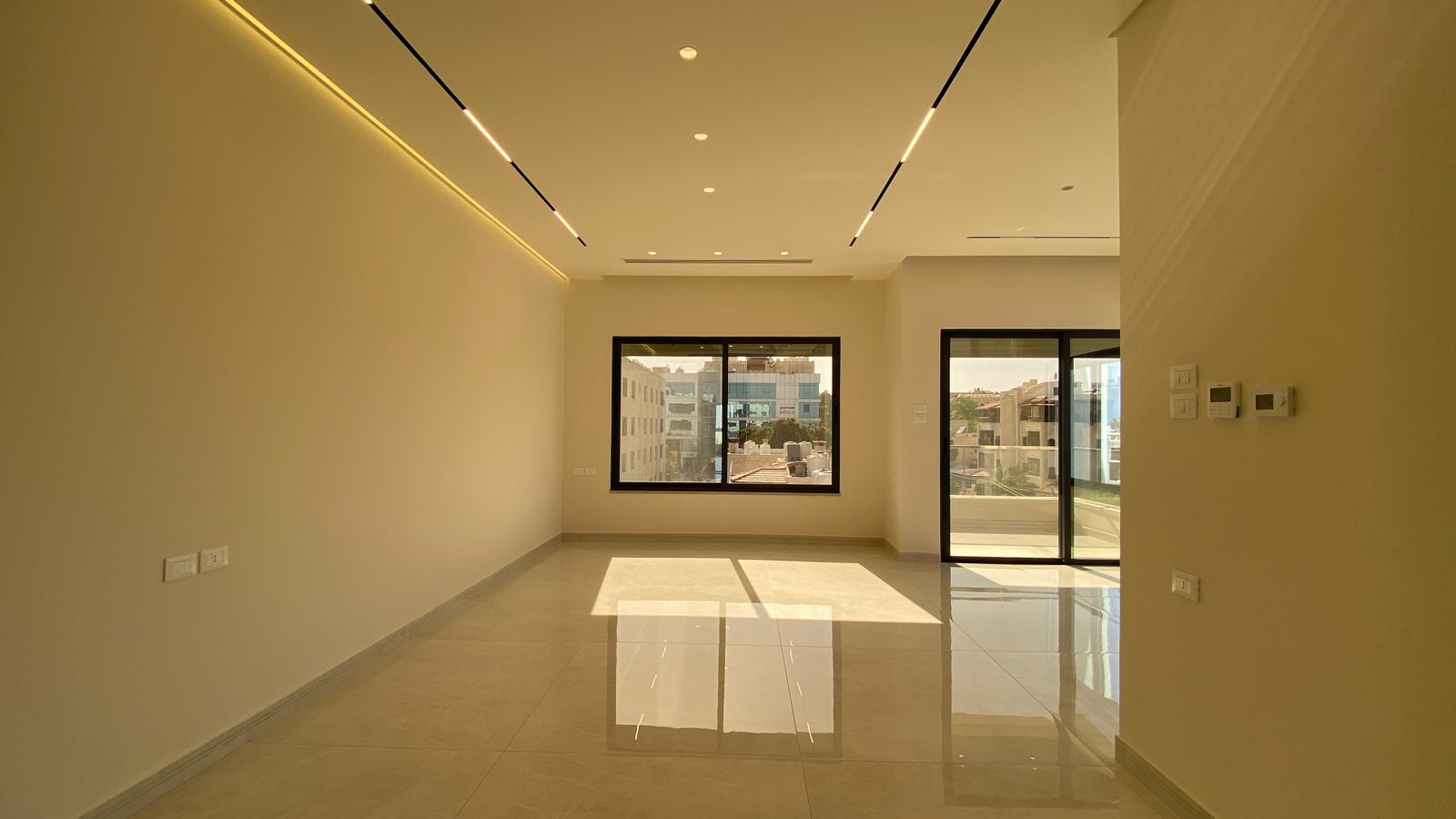 86cb5836-a70e-4de6-9c24-169a451fa0cb Bright empty living room with large windows and a sliding glass door to a balcony, sunlit glossy tile floor.
