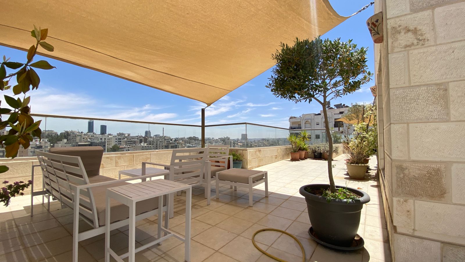 87989e08-6b2e-41ec-9917-b1ed894c254e Sunny rooftop terrace with white patio furniture, beige shade sail, and potted plants overlooking a city skyline.