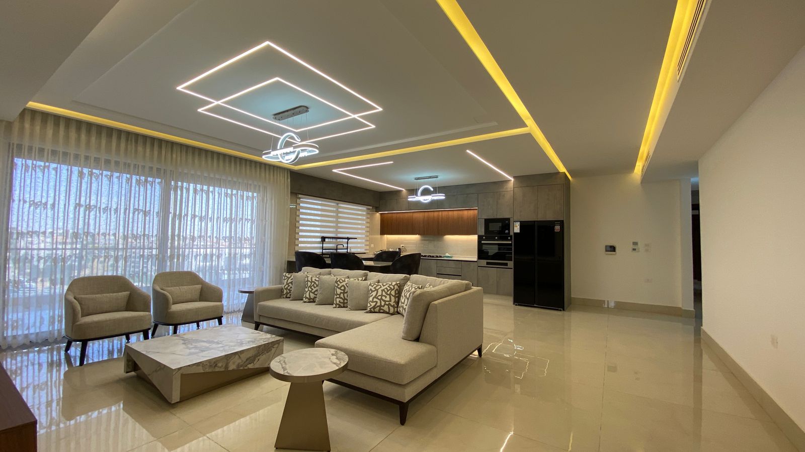 87d77df2-0f59-4a59-b52f-d14892821545 Open-concept living room with a beige L-shaped sofa, two armchairs, and a marble coffee table, kitchen in the background.