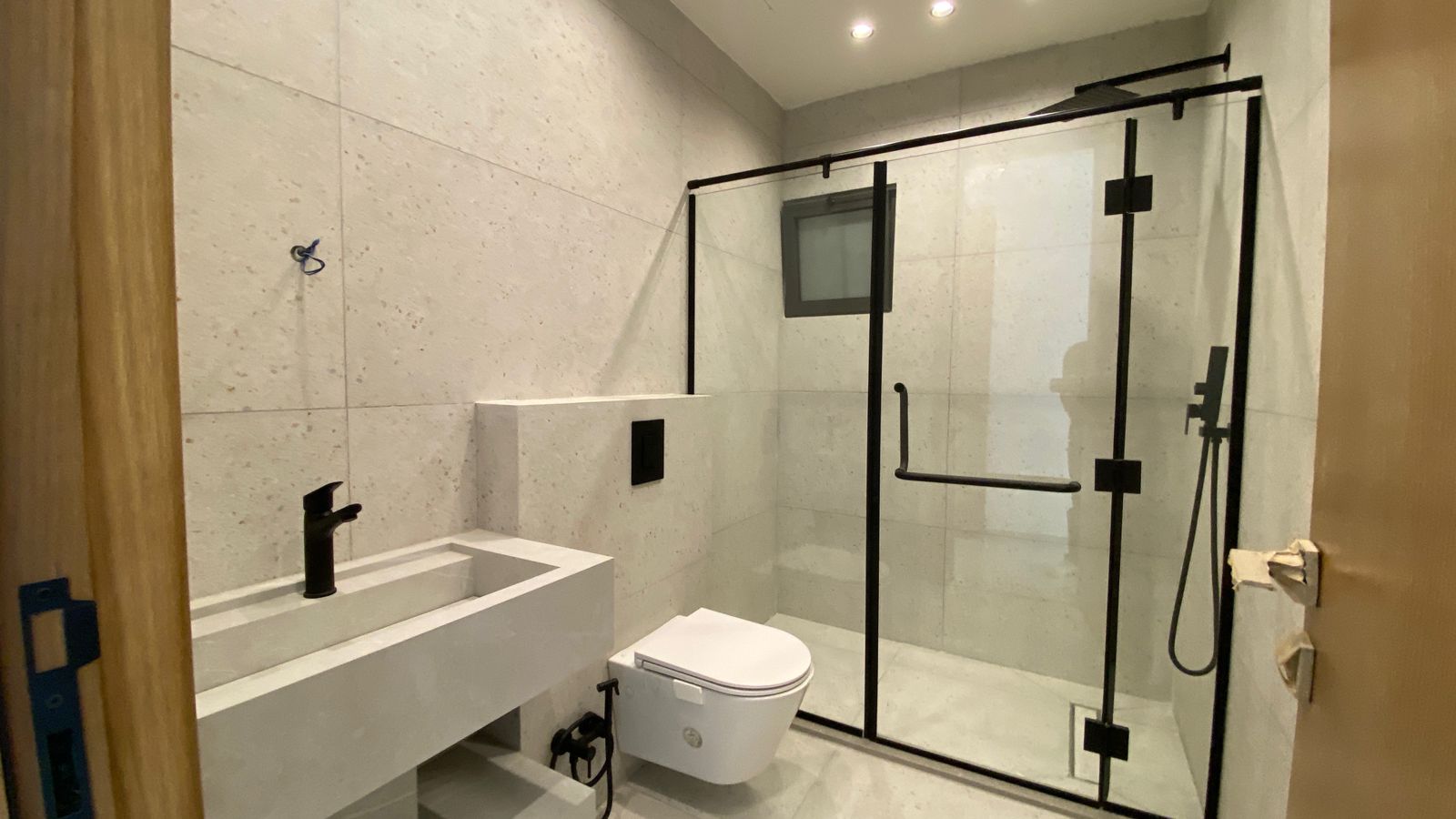 886bb660-fe63-4cdd-a4ef-2cd23fb5716f Modern bathroom featuring a glass shower enclosure with black metal frame, wall-mounted toilet, and concrete-style sink with black faucet.