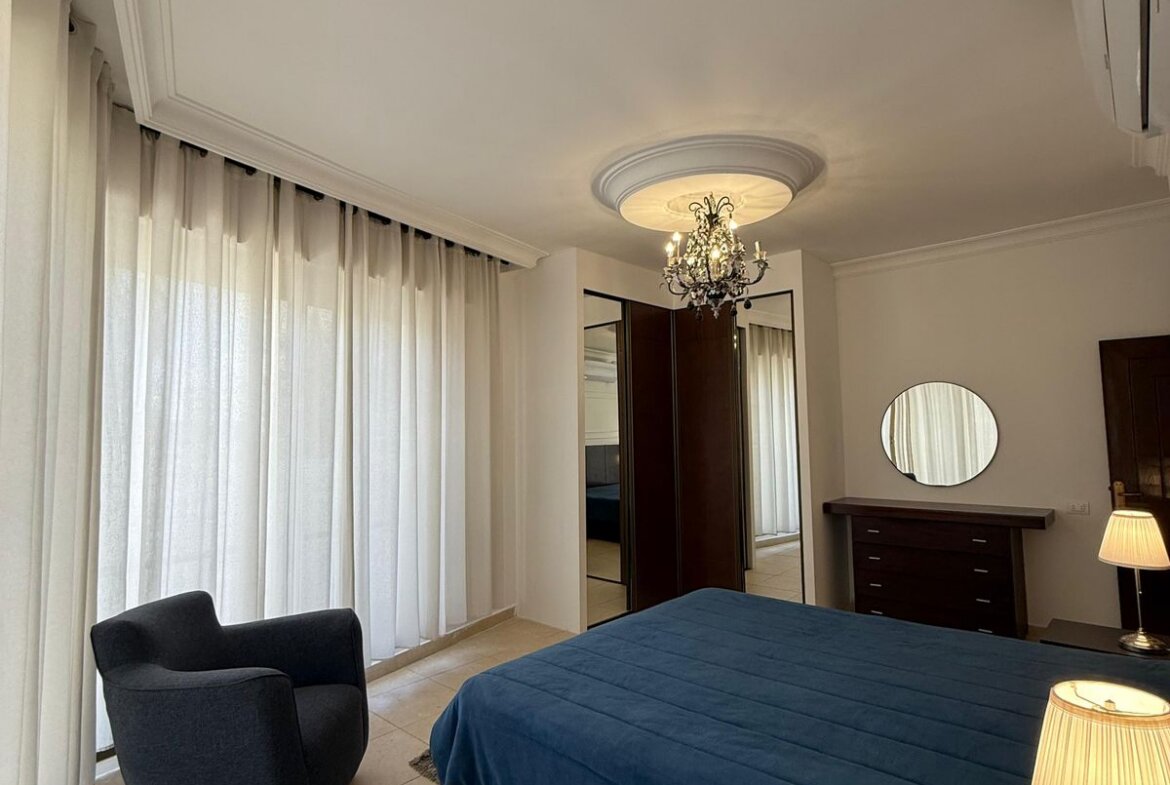 Bedroom with a large bed covered in a dark blue quilt, dark wood dresser with a round mirror, and a crystal chandelier on the ceiling medallion.