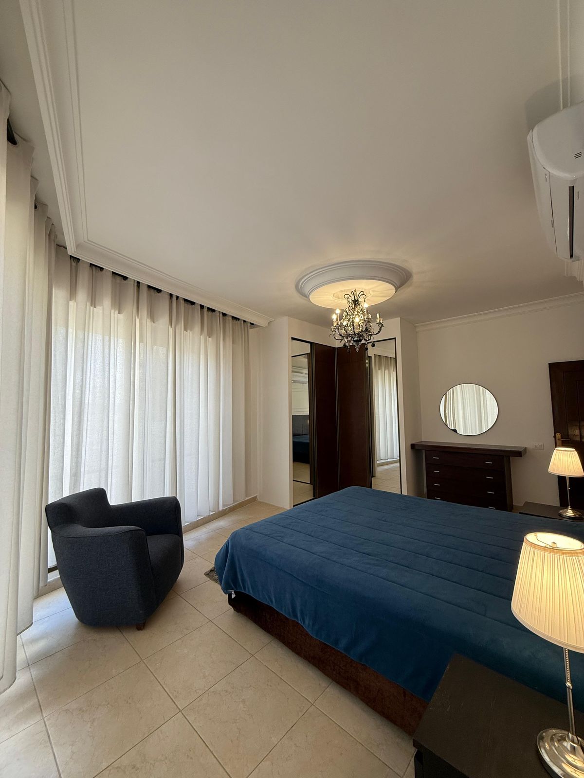 Bedroom with a large bed covered in a dark blue quilt, dark wood dresser with a round mirror, and a crystal chandelier on the ceiling medallion.