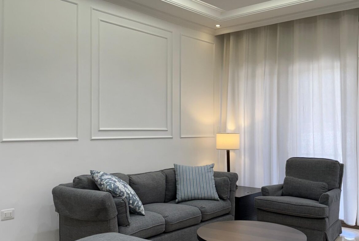Modern living room with gray sofa set, matching armchair and ottoman, circular coffee table, and light-filled windows with sheer curtains.