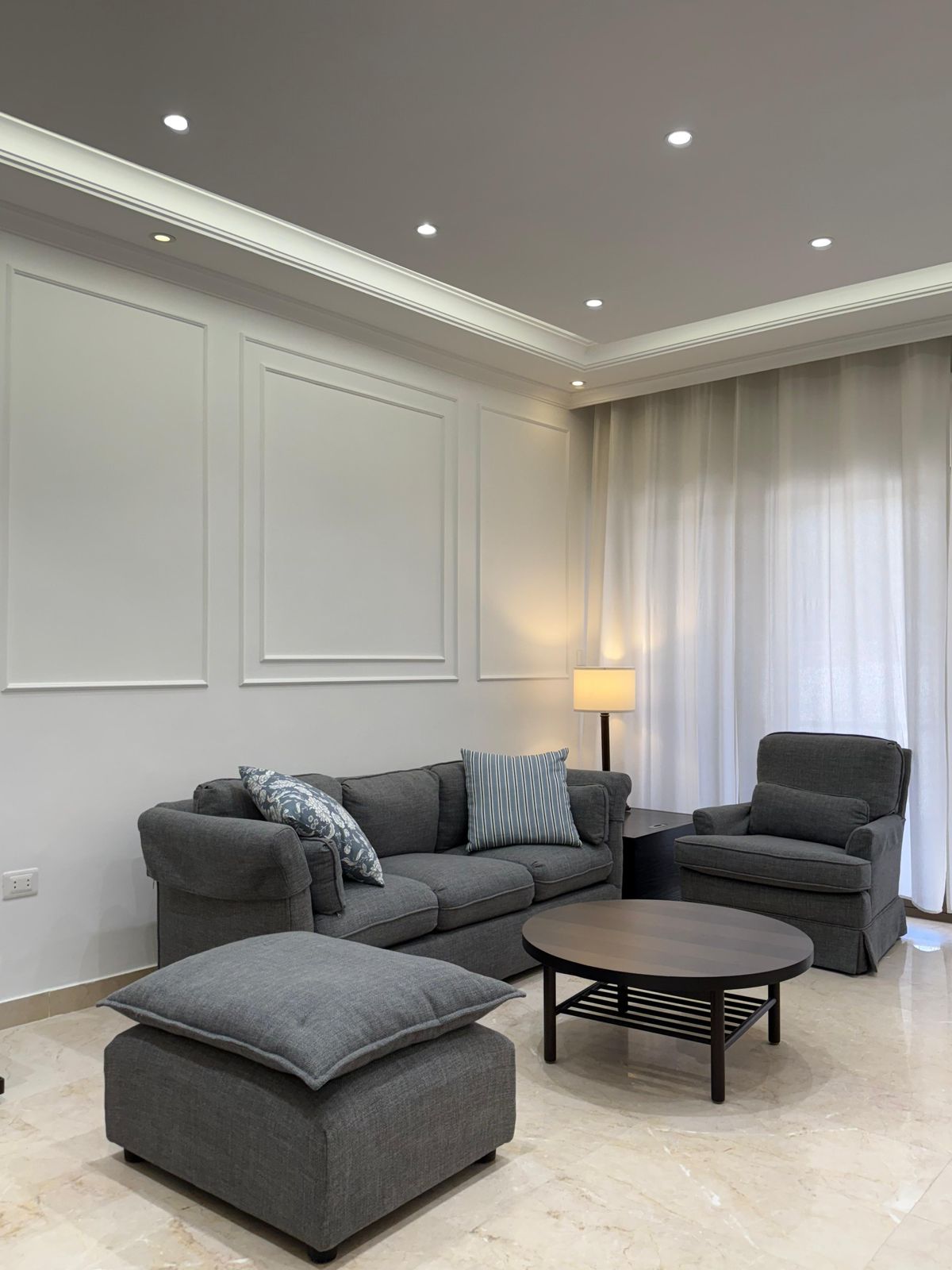 Modern living room with gray sofa set, matching armchair and ottoman, circular coffee table, and light-filled windows with sheer curtains.