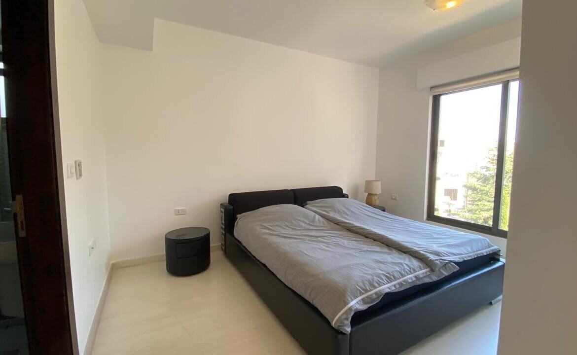 89b01837-0315-4e0b-8099-63e153546fc6 Bright minimalist bedroom with a dark low bed, two gray duvets, a round black nightstand, and a large window letting in daylight.