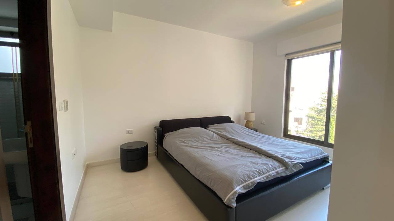 89b01837-0315-4e0b-8099-63e153546fc6 Bright minimalist bedroom with a dark low bed, two gray duvets, a round black nightstand, and a large window letting in daylight.