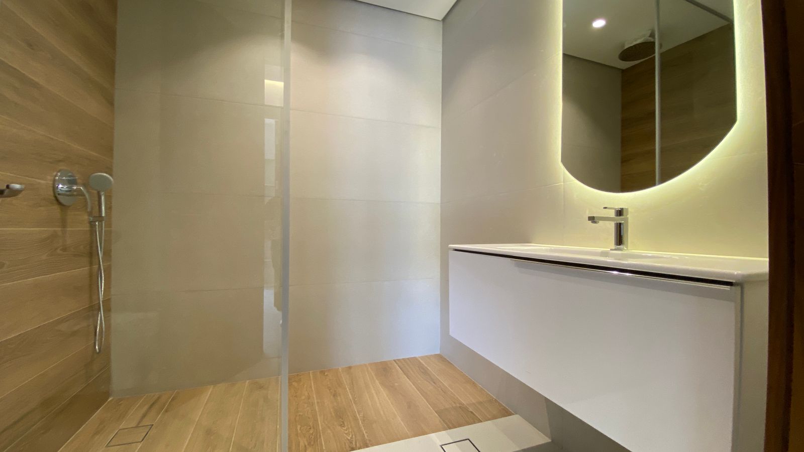 89dbae04-b70a-4e85-81af-a8693f77659d Modern bathroom with a wooden-floor shower area, glass divider, and a white floating vanity with a backlit, rounded mirror and chrome faucet.