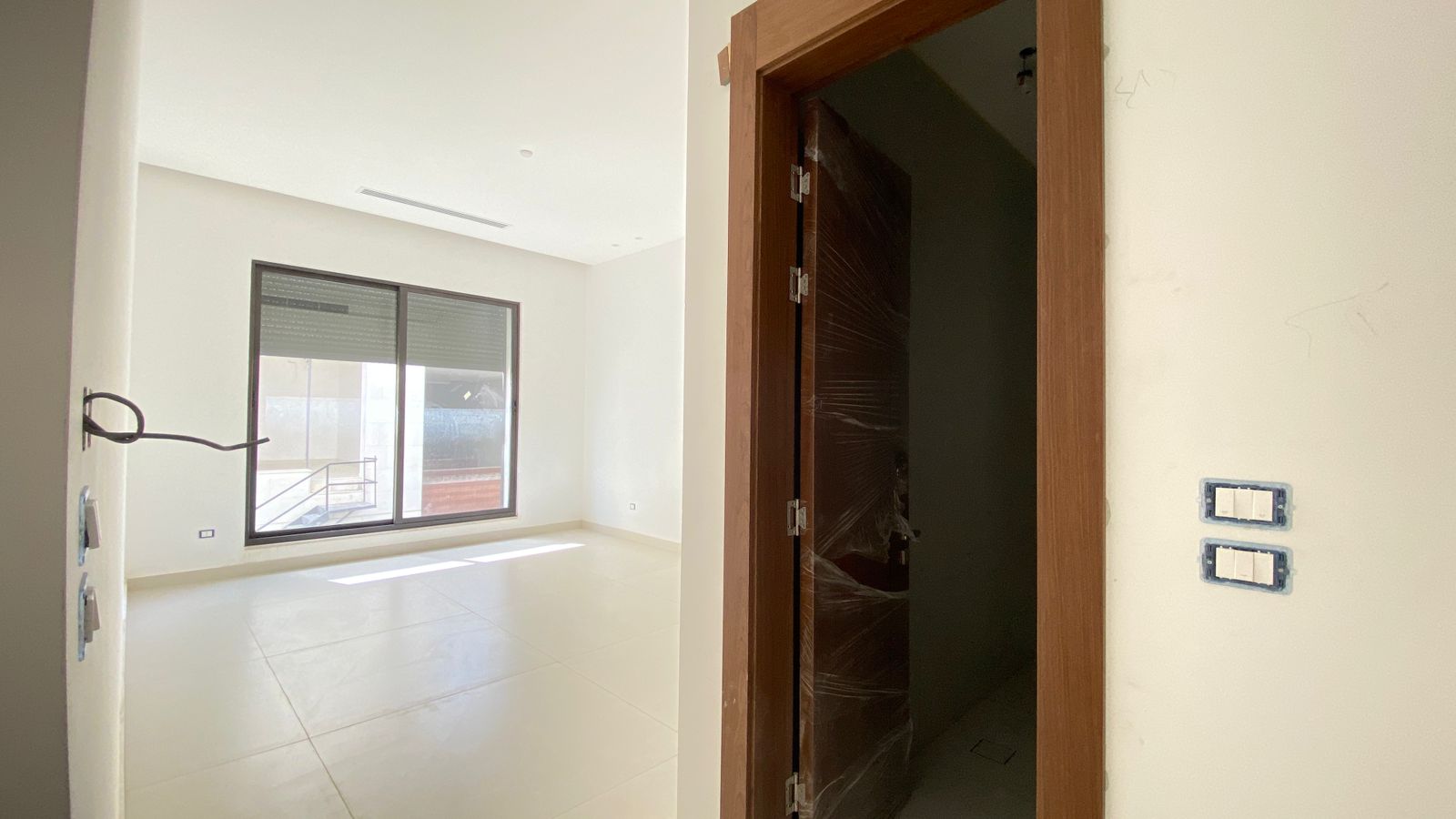 8a91b368-8eb8-46a5-b833-595cbdc1f969 Empty bright room with a large window/sliding door, white glossy floor tiles, and a wooden-framed doorway to a dark hallway.